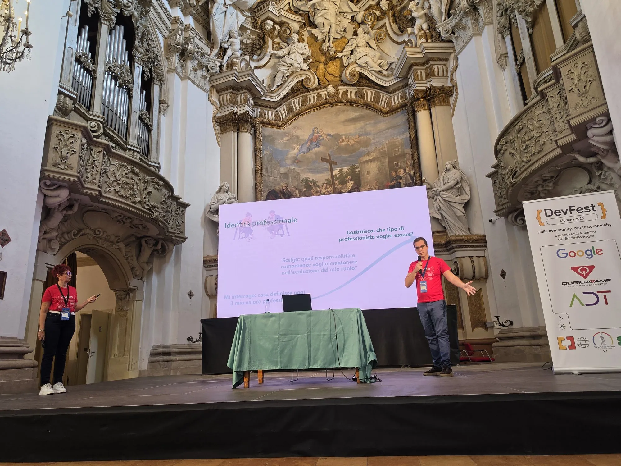 Isabella Flecchia e Gaetano Castaldo sul palco del DevFest Modena 2026, nella chiesa della Fondazione San Carlo, davanti alla slide con le tre domande sull’identità professionale